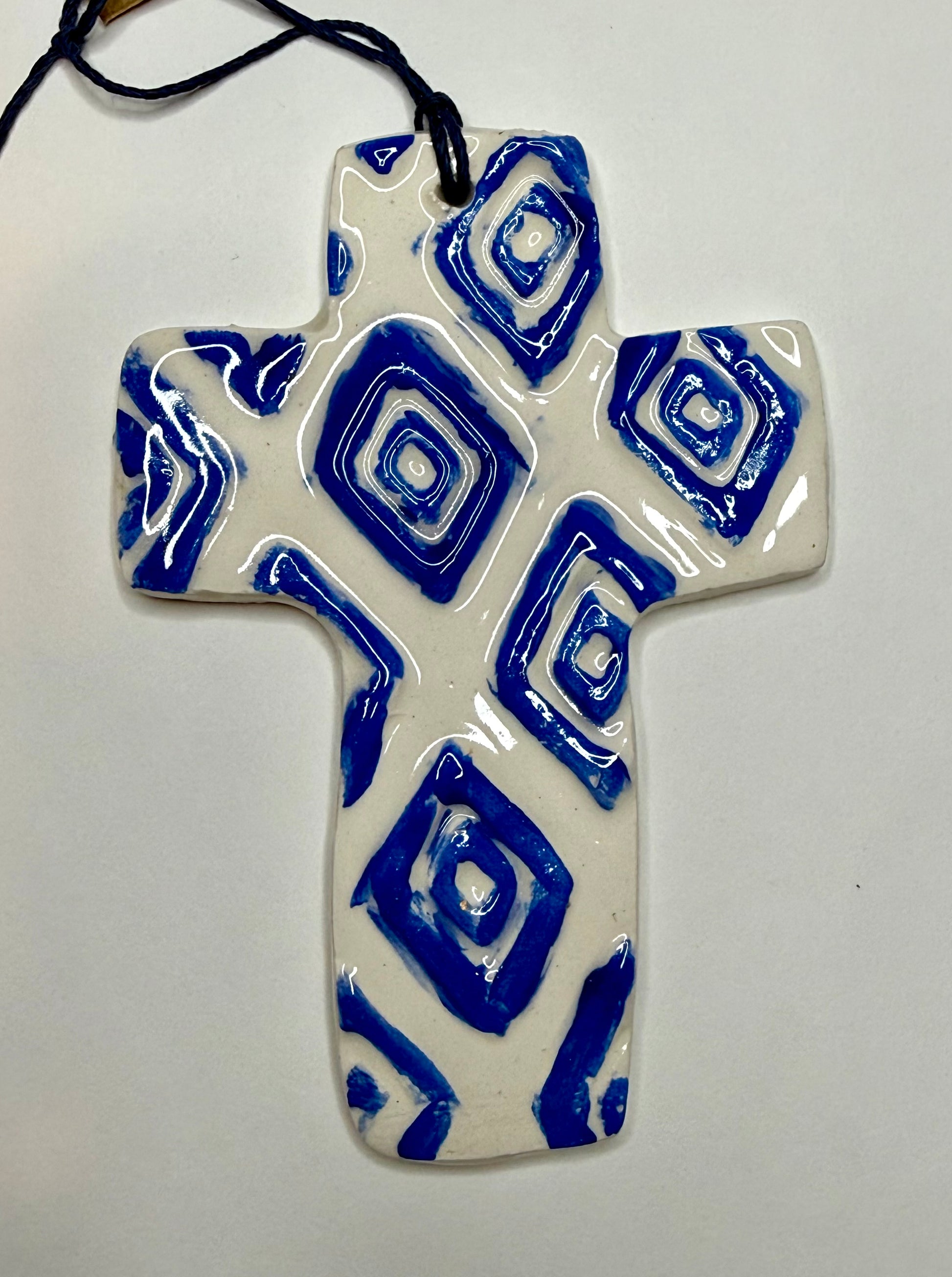 White and blue ceramic cross by Lorena Williams, featuring intricate designs and a glossy finish on a plain background.